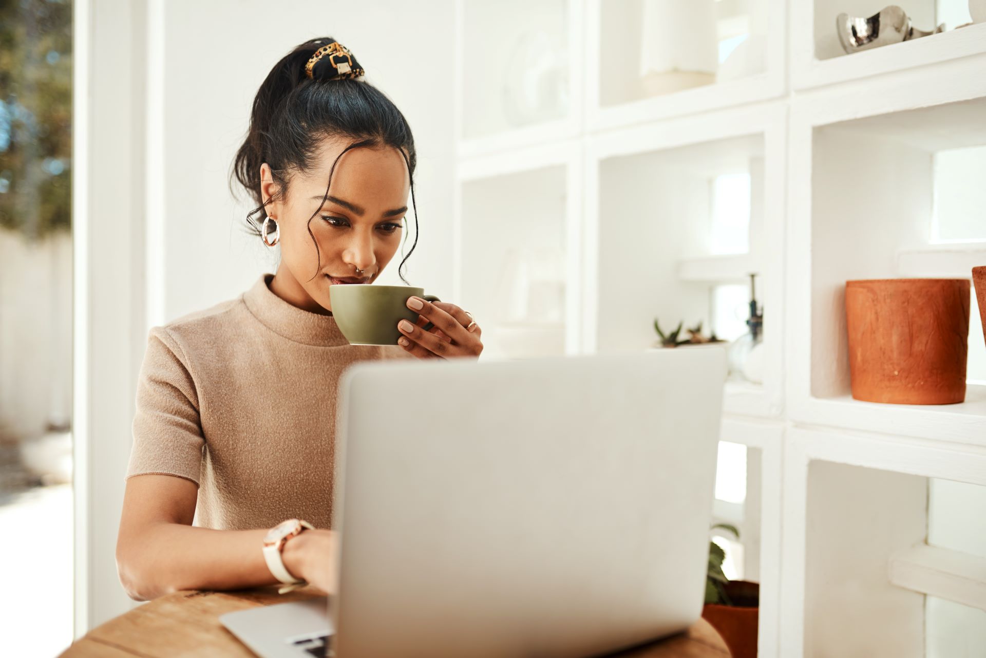 Junge Frau trinkt Kaffee und arbeitet konzentriert an einem Laptop in einem stilvollen, hellen Raum.